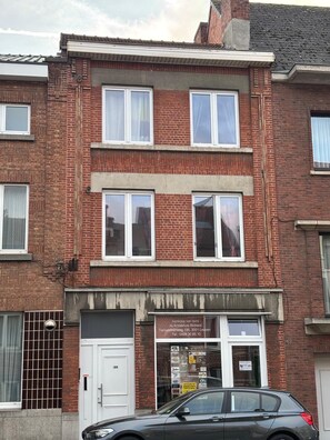 A two-story brick building with a ground-floor shop and a car parked in front. - Adorable apartment in nice Leuven perfect for your city stay (Leuven)