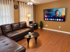 A living room with a leather sofa, a coffee table, a TV mounted on the wall displaying a movie, and a clock on the wall.