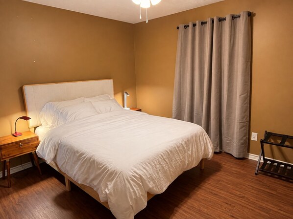 A neatly made bed in a room with a wooden floor, a nightstand with a lamp, and a window with curtains.