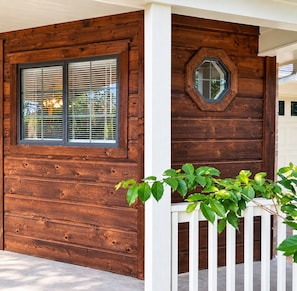 A wooden cabin with a window and a door, a white fence, and a plant in the foreground.