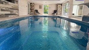 A modern indoor swimming pool with clear blue water, surrounded by tiled walls and a glass door leading outside. - Mereside House | Heated Indoor Pool, Hot Tub & Cinema (Allostock)