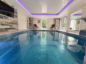 En moderne indendørs swimmingpool med krystalklart blåt vand, omgivet af fliserede vægge og en glasdør til terrassen.