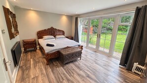A bedroom with a large bed, a wooden nightstand, a dresser, and a sliding glass door leading to a garden. - Mereside House | Heated Indoor Pool, Hot Tub & Cinema (Allostock)
