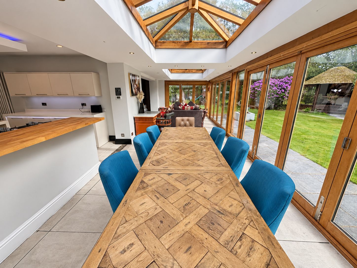 A modern kitchen with a long wooden table and blue chairs, a dining area with a view of the garden through large glass doors.