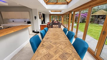 A modern kitchen with a long wooden table and blue chairs, a dining area with a view of the garden through large glass doors.
