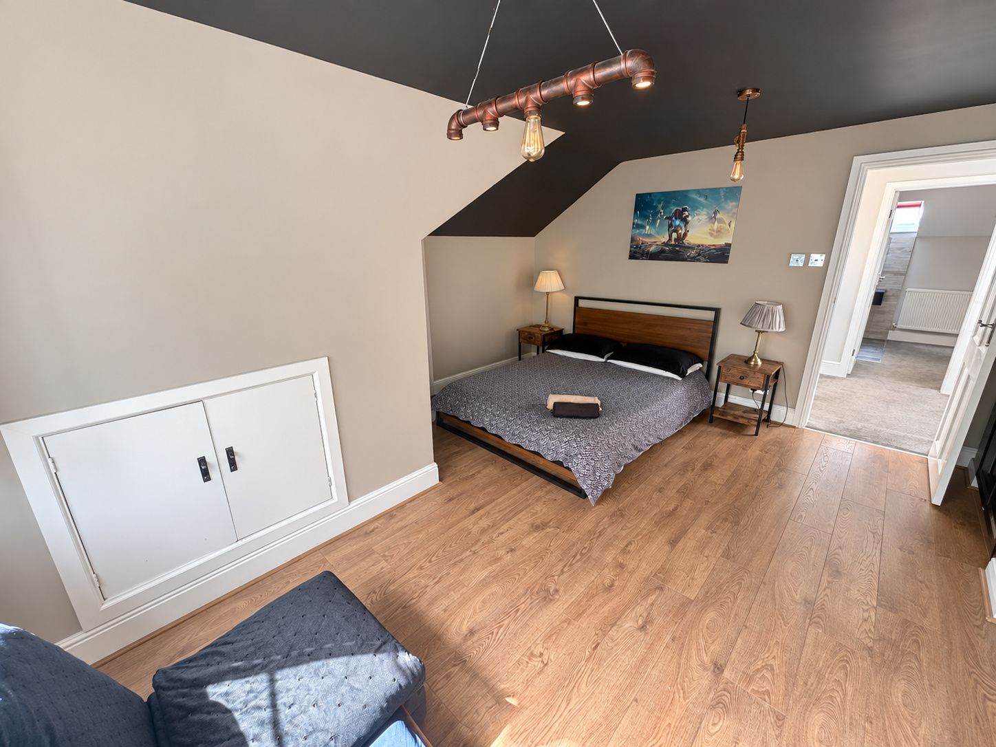 A bedroom with a sloped ceiling, a bed with a patterned bedspread, a bedside table with a lamp, and a wall-mounted picture.