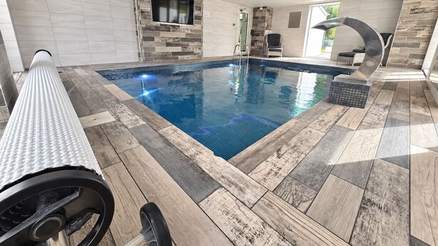 A modern indoor swimming pool with a wooden floor, a built-in TV, and a seating area with a curved bench. - Mereside House | Heated Indoor Pool, Hot Tub & Cinema (Allostock)
