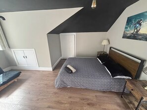 A bedroom with a sloped ceiling, a bed with a patterned bedspread, a nightstand with a lamp, a wooden bench, and a wardrobe.