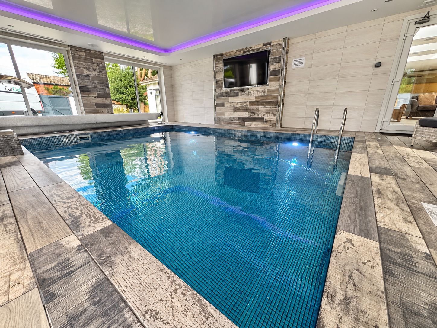 A swimming pool with a tiled edge, a built-in TV, and a wooden deck surrounding it.