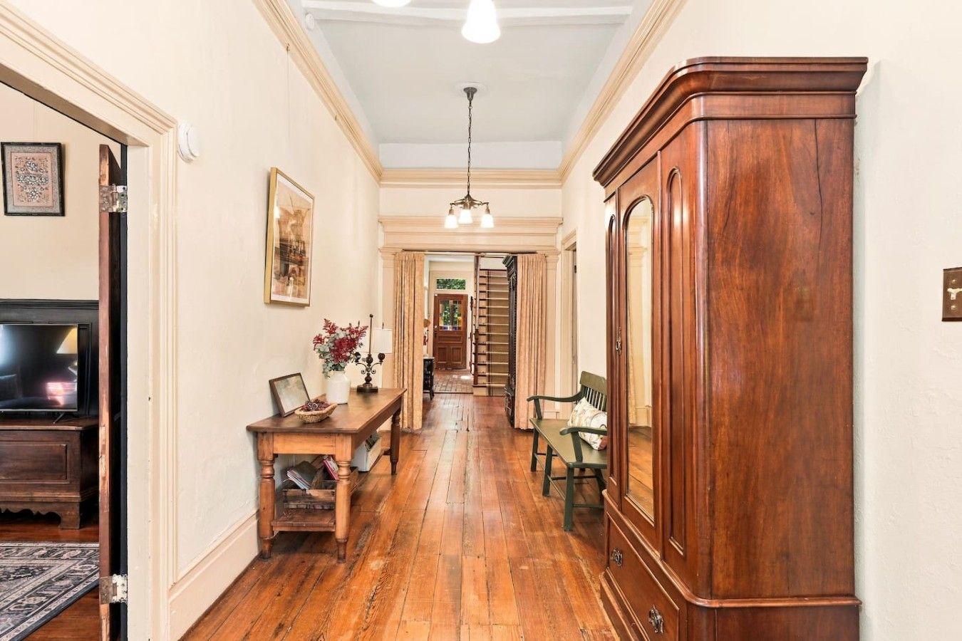 A hallway with a wooden floor, a wooden cabinet, a table with a vase, and a television.