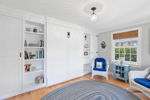 Ein Zimmer mit weißen Einbauschränken, einem Korbstuhl mit Kissen, einem blauen Sofa, einem gemusterten Teppich und einem Fenster mit Blick auf Grünflächen.
