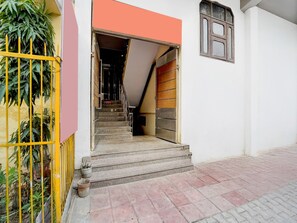 L’entrée d’un hôtel, avec un escalier menant à un palier, un espace clos agrémenté de plantes et une fenêtre dotée d’un volet en bois.