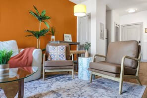 Un salon avec un canapé, un fauteuil et une table basse en verre. On y trouve des plantes en pot et un tapis sur le sol.