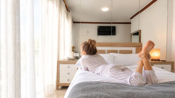 Ein Schlafzimmer mit einem Bett, einem Nachttisch, einer Lampe, einem an der Wand befestigten Fernseher und einem Fenster mit Vorhängen.