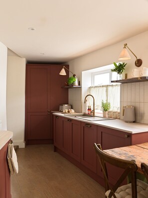 A kitchen with wooden cabinets, a countertop, a sink, a window with curtains, and a hanging lamp. - Charming Two Bedroom Cottage in Malmesbury (Malmesbury)