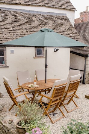 A patio with a round wooden table and six chairs, a green umbrella, and a potted plant. - Charming Two Bedroom Cottage in Malmesbury (Malmesbury)