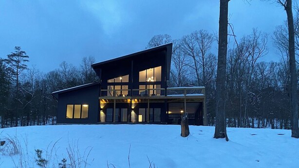 Une maison moderne à deux étages, dotée de grandes fenêtres et d’un balcon, entourée d’un paysage enneigé et d’arbres.