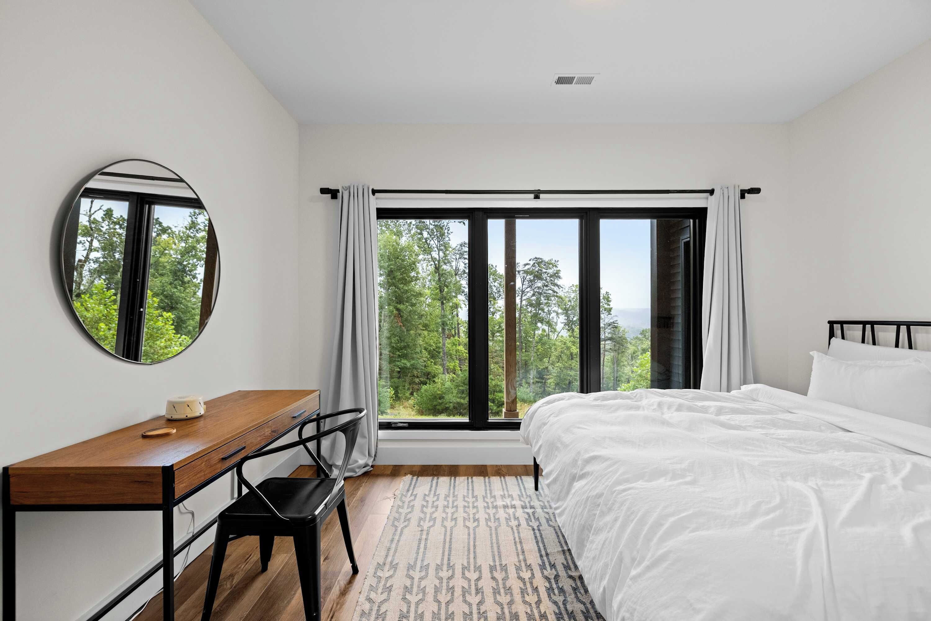 Quarto com cama grande, escrivaninha, cadeira, espelho redondo e vista para árvores pela janela.