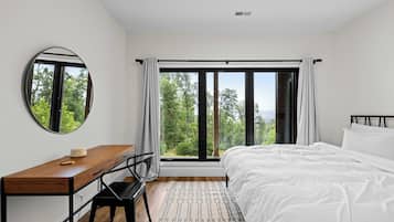 Una camera da letto con un letto grande, una scrivania, una sedia, uno specchio rotondo e vista sugli alberi dalla finestra.