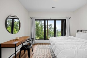 A bedroom with a large bed, a desk, a chair, a round mirror, and a view of trees through the window.