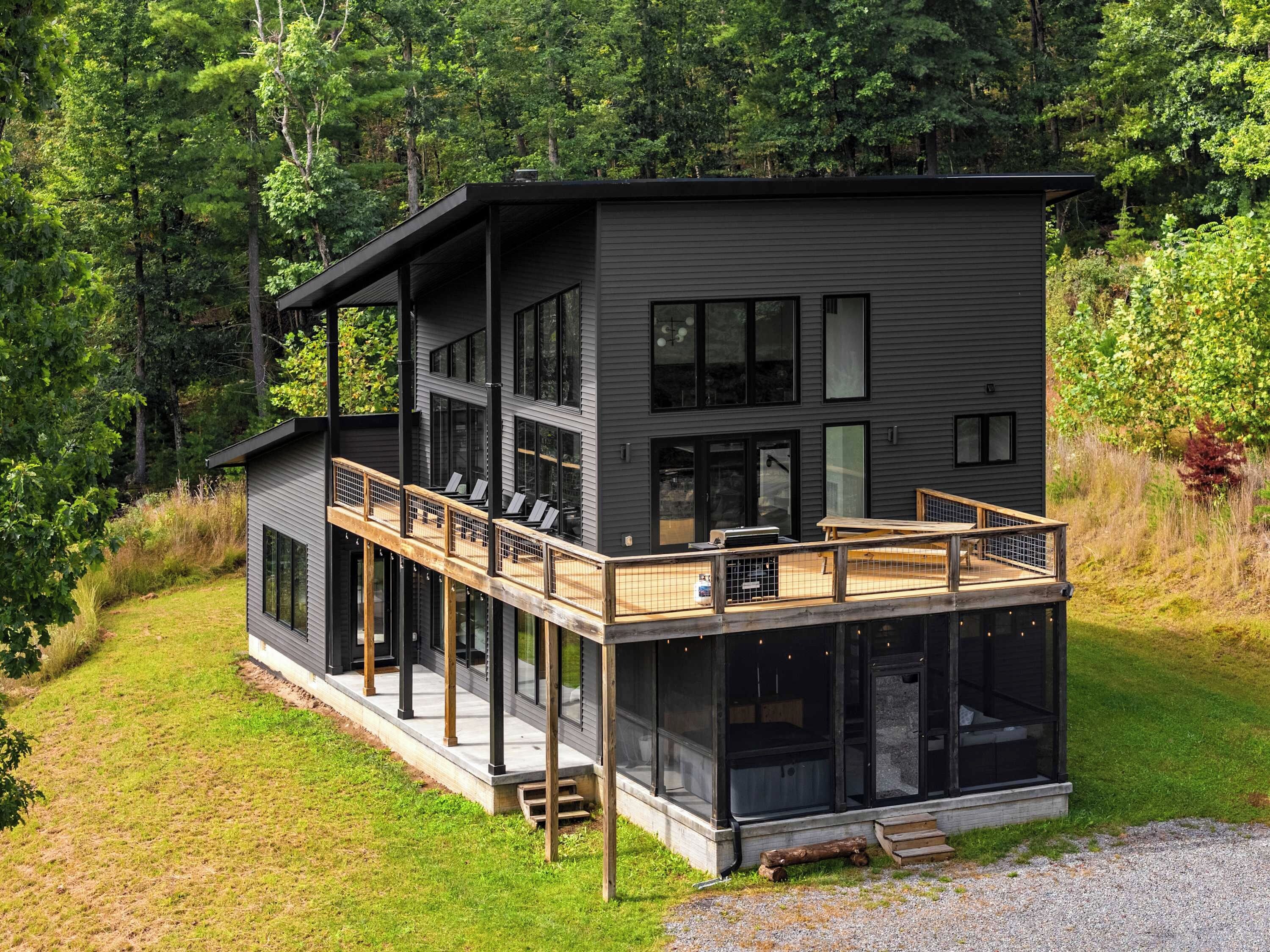 A modern two-story house with a deck, large windows, and a natural forest backdrop.