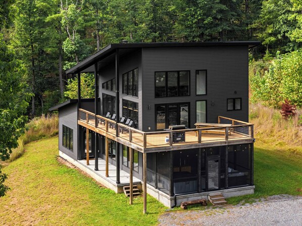 A modern two-story house with a deck, large windows, and a natural forest backdrop.