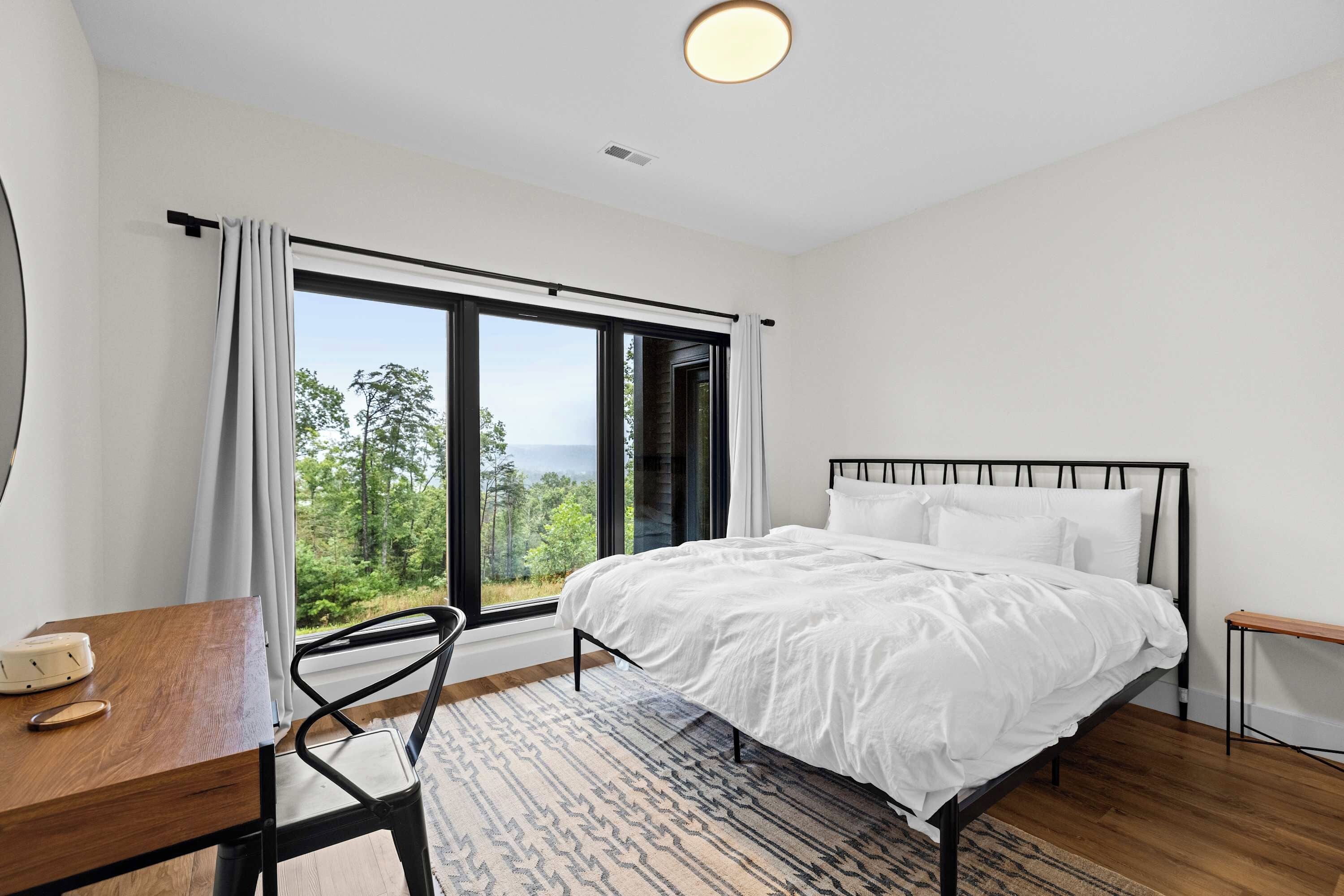 A bedroom with a bed, a desk, and a chair. There is a large window with a view of trees and the sky.