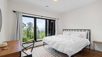 A bedroom with a bed, a desk, and a chair. There is a large window with a view of trees and the sky.