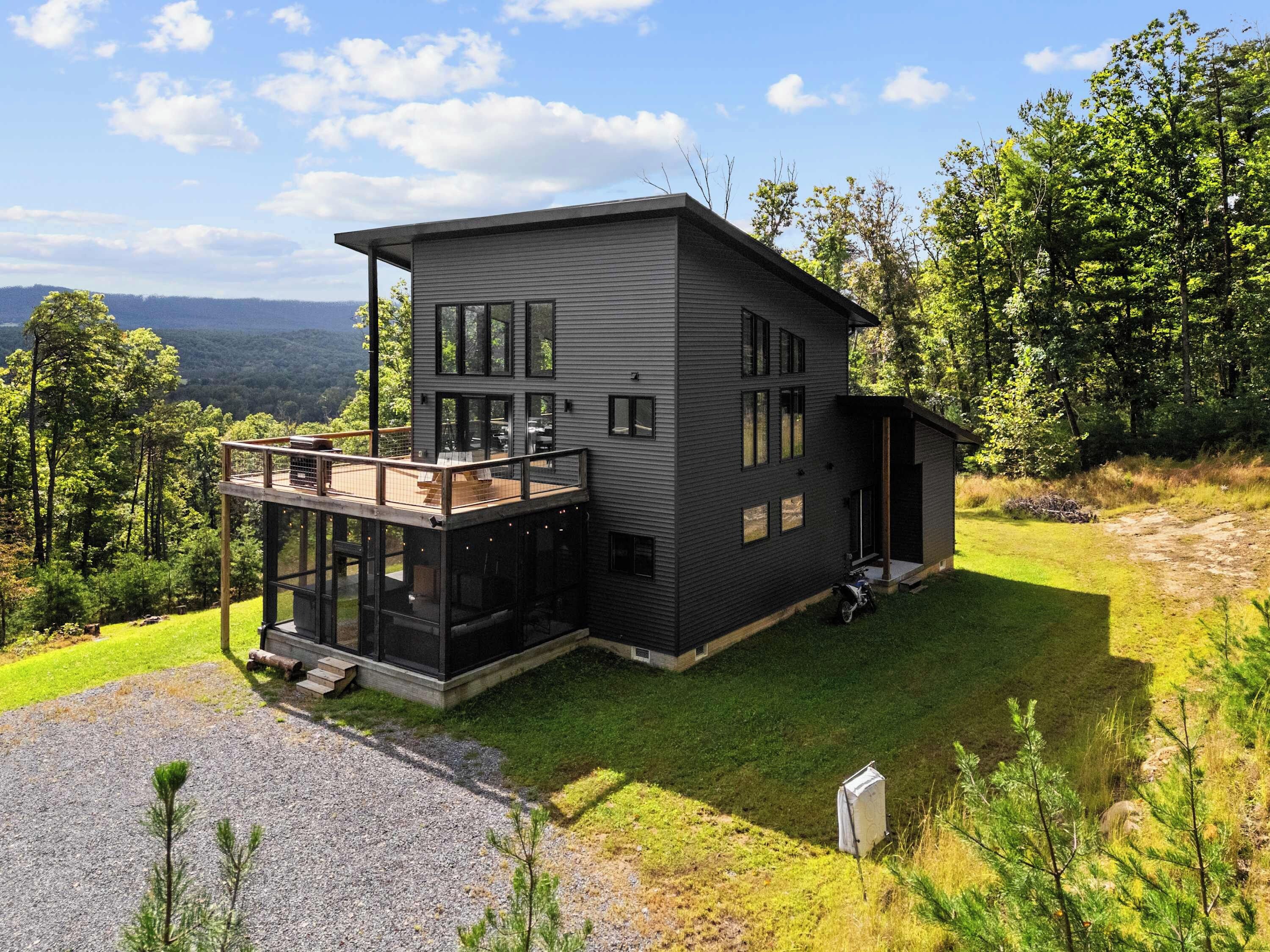 A modern two-story house with a deck, large windows, and a view of the surrounding forest.