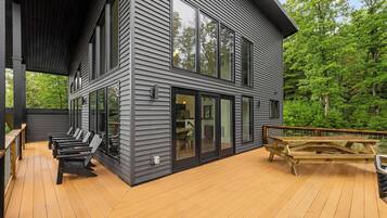 A modern two-story house with a deck, large windows, and a view of a forest.