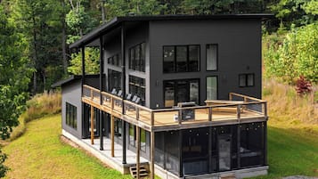 Ein modernes, zweistöckiges Haus mit einer Terrasse, großen Fenstern und einem Wald im Hintergrund.