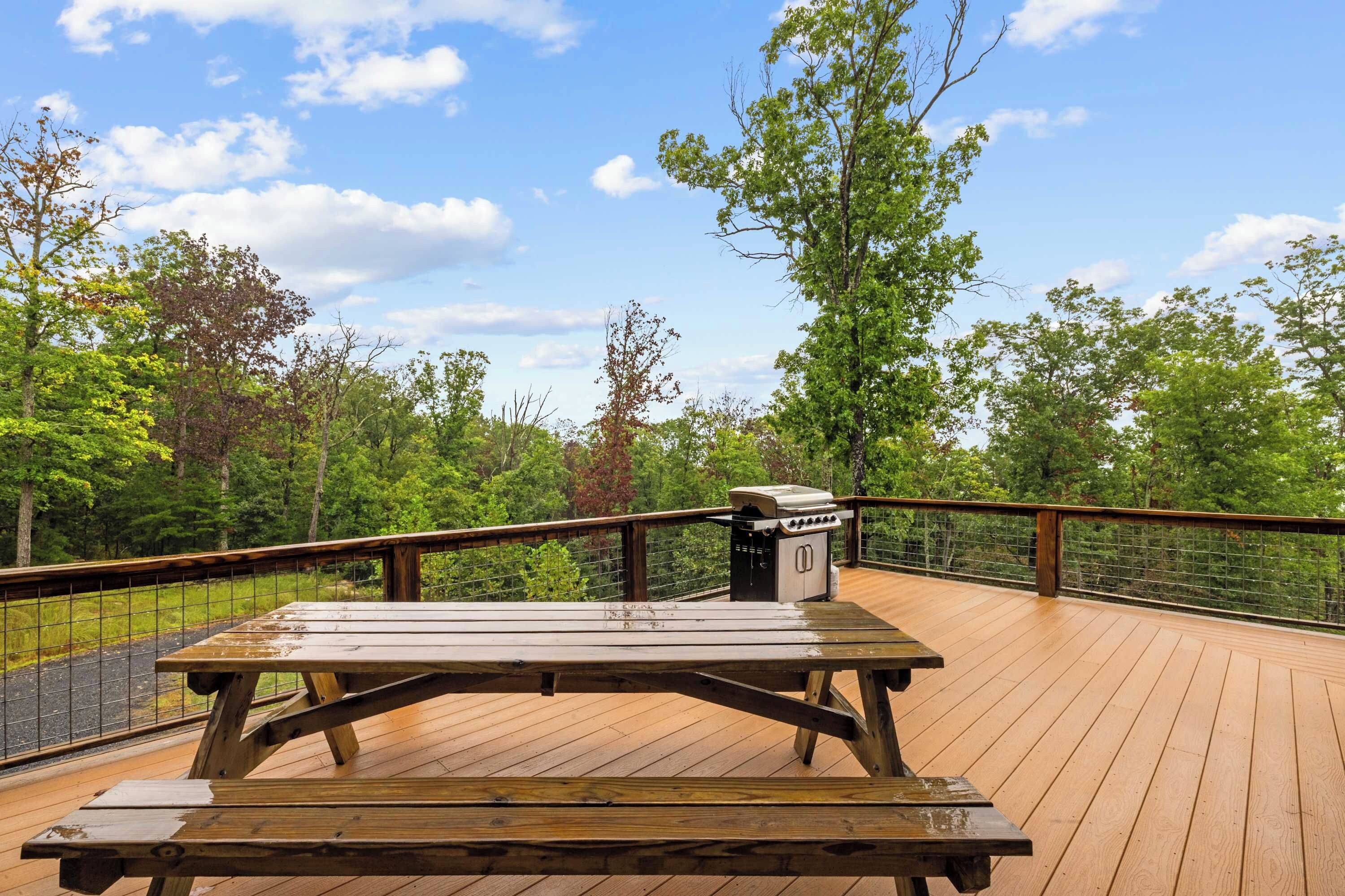 A wooden deck with a picnic table and grill, surrounded by trees and a clear sky.