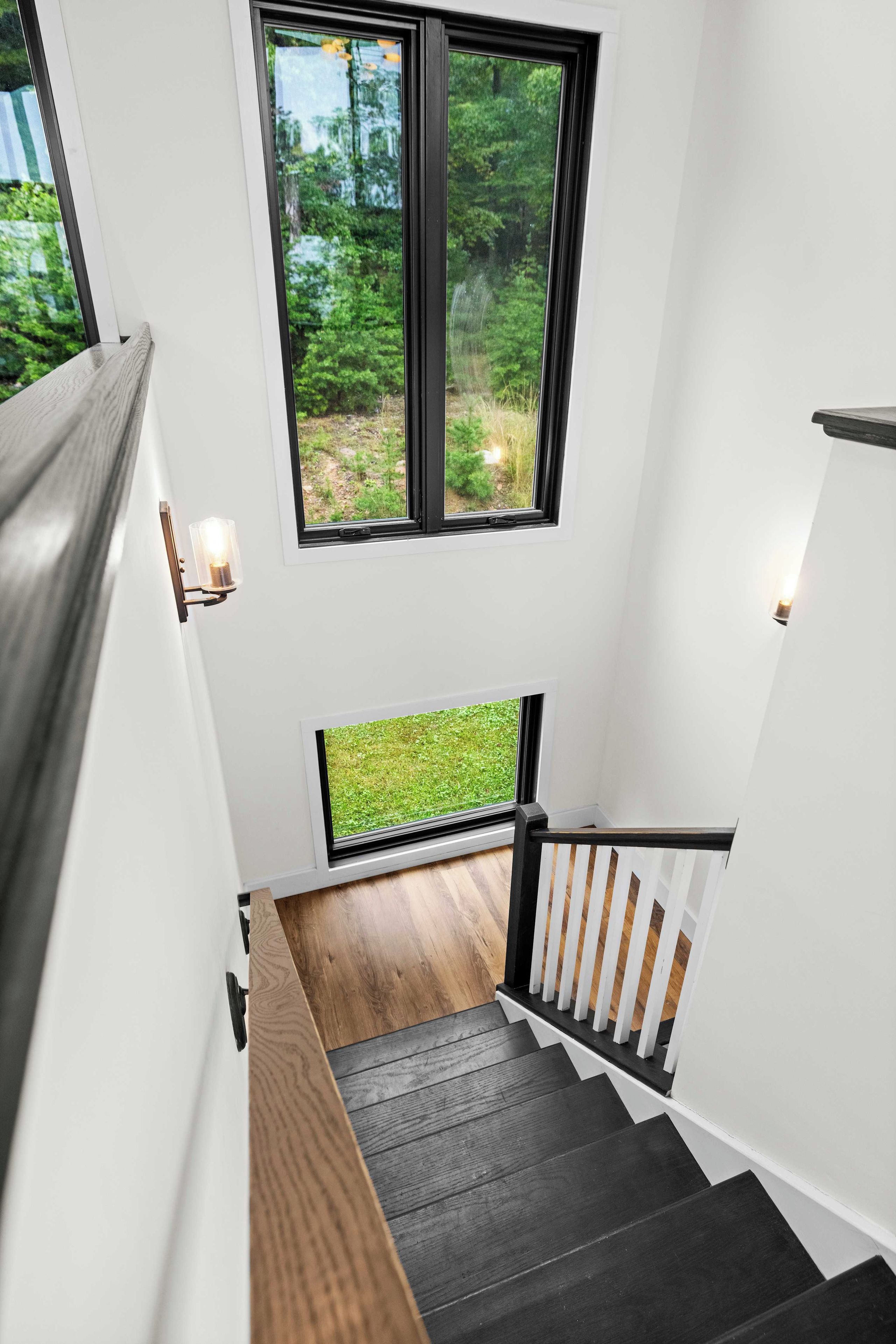 A modern staircase with a wooden handrail and a view of a green outdoor area through large windows.