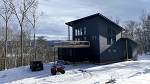 Une maison moderne à deux étages, dotée d’une terrasse couverte, entourée d’un paysage enneigé et d’arbres.