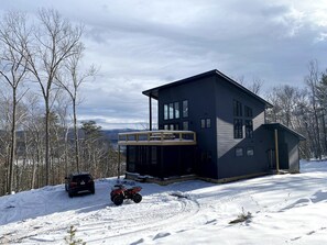 Uma casa moderna de dois andares com varanda coberta, cercada por uma paisagem nevada e árvores.