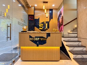 A hotel reception desk with a check-in/out time display, a reception area, and a staircase.
