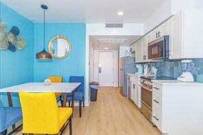 A compact kitchen with white cabinets, a microwave, a stove, and a sink. There is a dining area with a table and chairs, and a trash bin is visible. - Endless Ocean Views at Yachtsman S904 (Myrtle Beach)