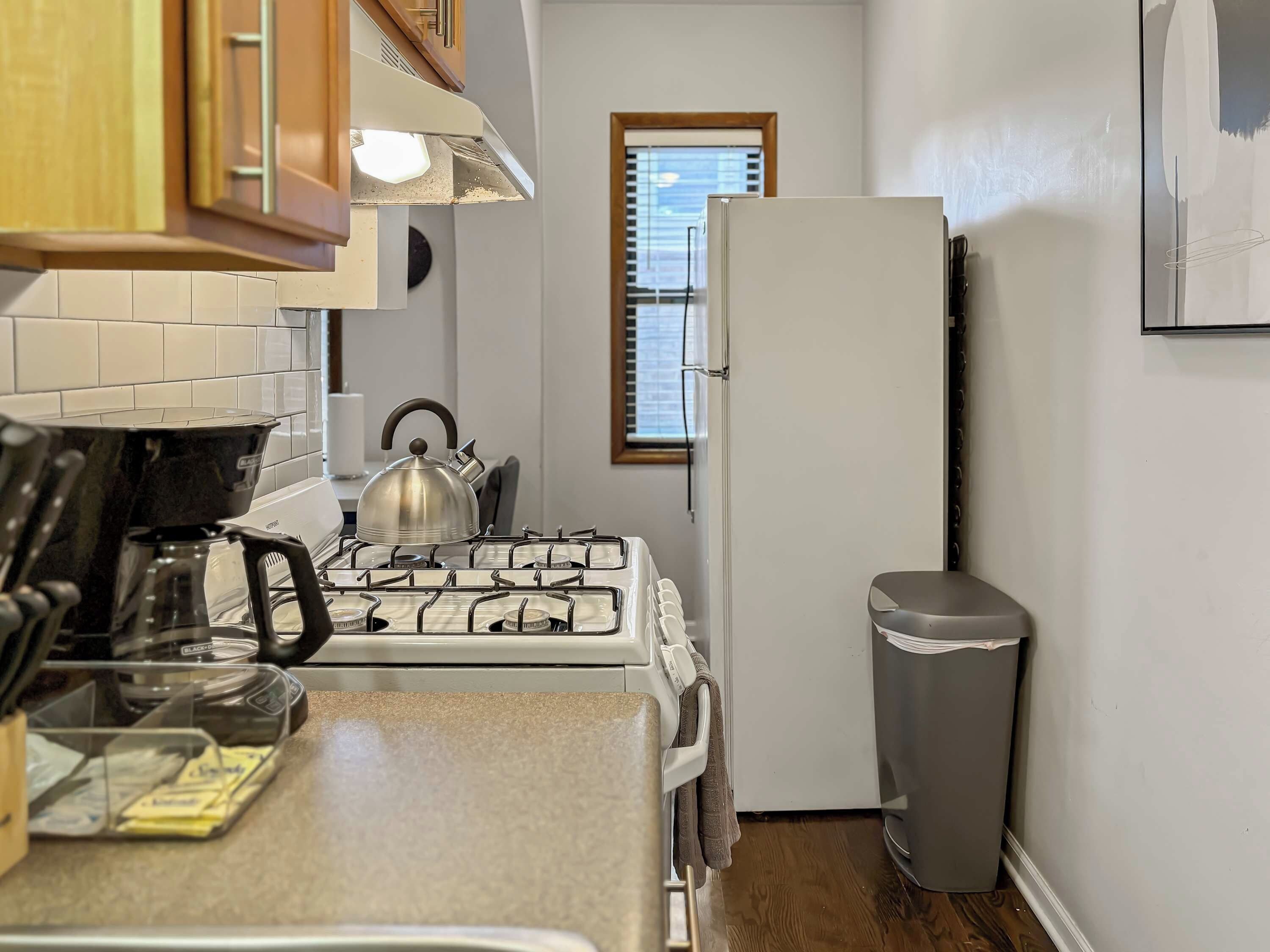 A compact kitchen with a gas stove, kettle, and a trash bin.
