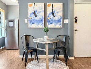 Un área de comedor con una mesa redonda, dos sillas y una planta pequeña. Hay dos cuadros abstractos en la pared sobre la mesa.