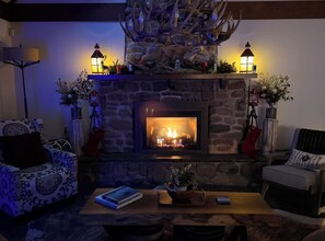 Un salon chaleureux avec une cheminée en pierre, deux fauteuils, une table basse et des objets décoratifs, comme un lustre en forme de bois de cerf et des lanternes.