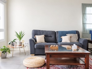 Ein modernes Wohnzimmer mit einem dunkelblauen Sofa, zwei weißen Kissen, einem Glastisch und einem gewebten Hocker. Übertöpfe und eine Lampe stehen auf einem Beistelltisch.