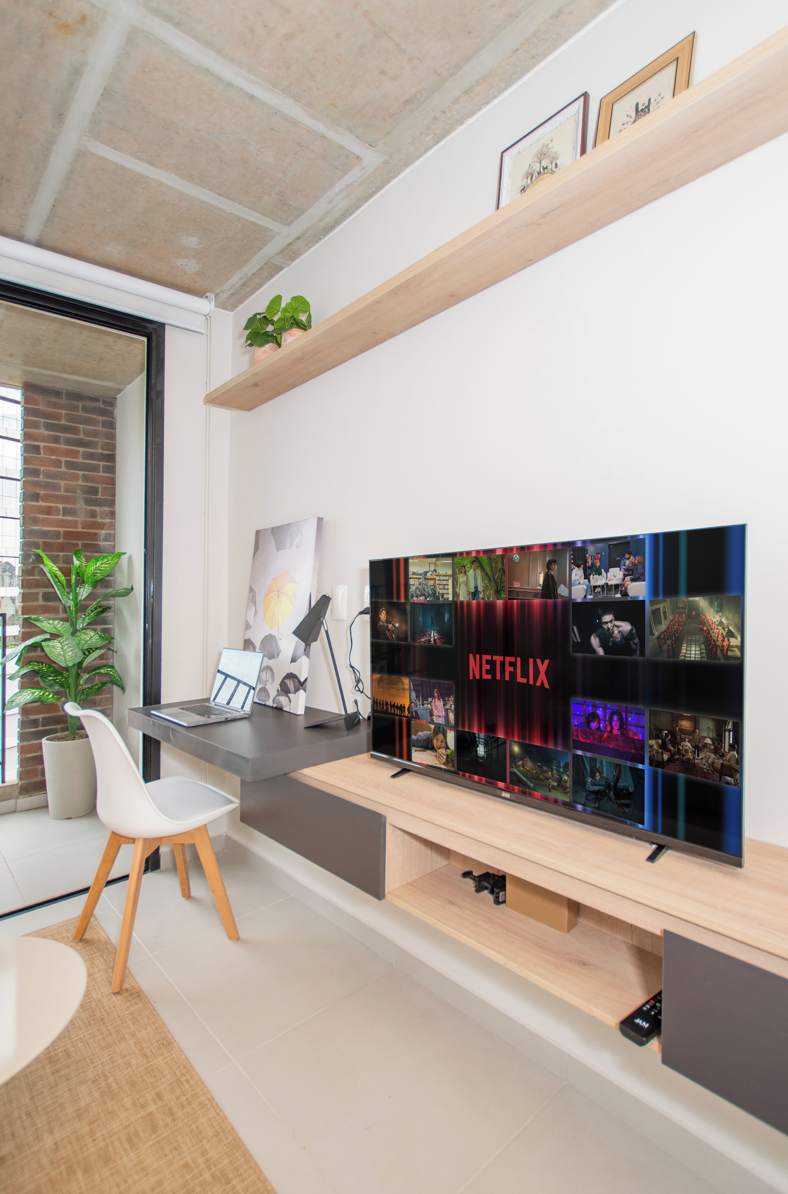 Una sala moderna con un televisor de pantalla plana grande montado en un estante de madera, una computadora portátil sobre un escritorio y una planta en maceta.