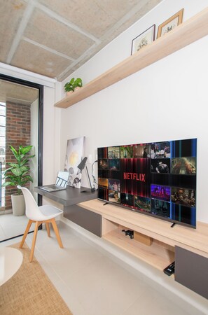 Una sala moderna con un televisor de pantalla plana grande montado en un estante de madera, una computadora portátil sobre un escritorio y una planta en maceta.