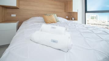 Una cama bien tendida con dos toallas dobladas, cabecera de madera, mesita de noche con lámpara y ventana con vistas.