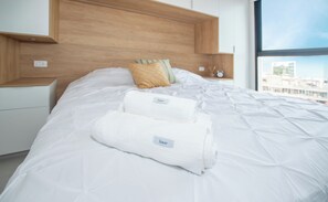 A neatly made bed with two folded towels, a wooden headboard, a nightstand with a lamp, and a window with a view. - Corporate Stays First del Sol - 707 (Asunción)