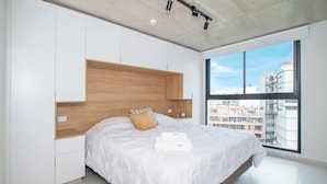 A modern bedroom with a large bed, built-in wooden headboard, white bedding, and a view of a cityscape through a large window. - Corporate Stays First del Sol - 707 (Asunción)