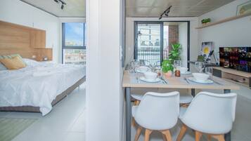 A modern bedroom with a large bed, a dining area with a table set for two, and a view of the city through a window.