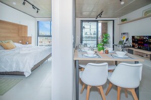 Une chambre moderne avec un grand lit, un coin repas avec une table dressée pour deux personnes, et une vue sur la ville par la fenêtre.