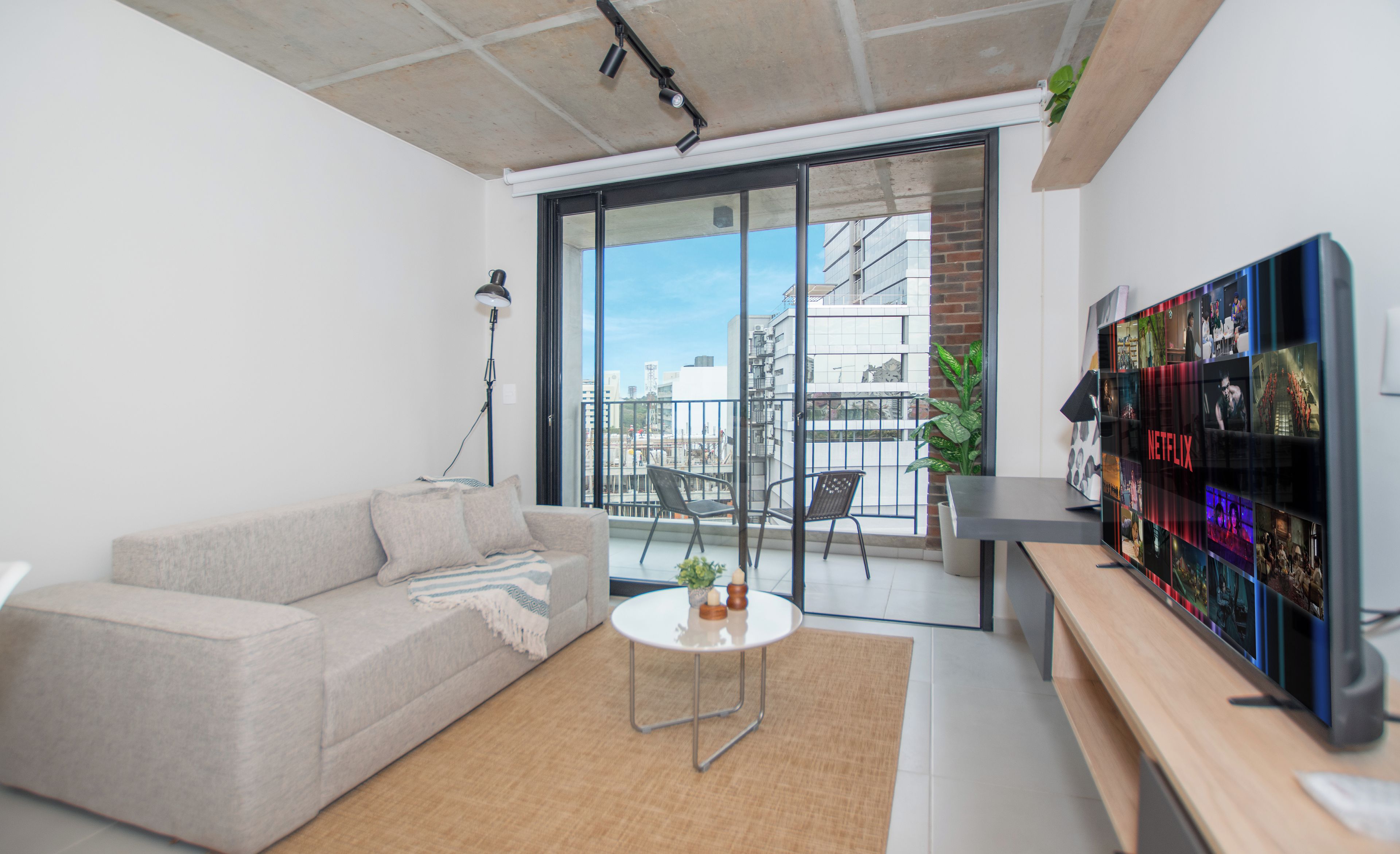 A modern living room with a sofa, a coffee table, a TV, and a balcony with a view of the city.