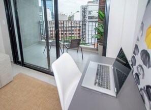 Un apartamento moderno con balcón, una computadora portátil sobre un escritorio y vistas panorámicas de la ciudad.